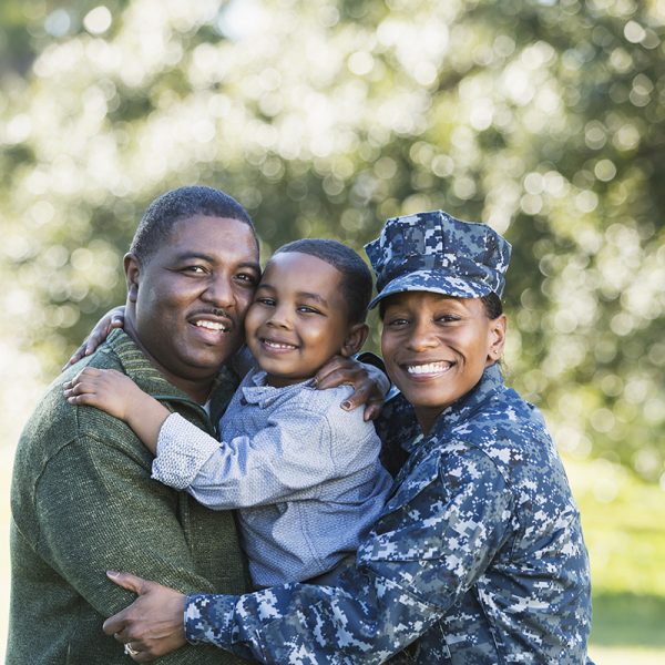 A military homecoming. Portrait of an African American woman wearing navy camouflage uniform standing outdoors with her family. Her husband is holding their 5 year old son who is in the middle between his parents, smiling at the camera. The main focus is on the woman.