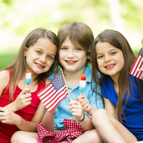 Three American children enjoy a summer picnic outdoors to celebrate USA's Memorial Day, Independence Day, or Labor Day holiday.  The three siblings enjoy yummy red, white, and blue popsicles. Two little girls and one little boy are all elementary ages. Beautiful nature background. American flags.