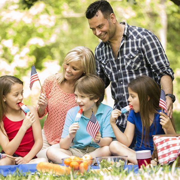 American family with three children enjoys a summer picnic outdoors to celebrate USA's Memorial Day, Independence Day, or Labor Day holiday.  The three children enjoy red, white, and blue popsicles. Father, mother. Two little girls and one little boy are elementary age. Beautiful nature background. American flags.