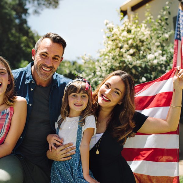 Happy family sitting together in their backyard holding the american flag behind them. Smiling couple with their kids celebrating american independence day holding american flag.